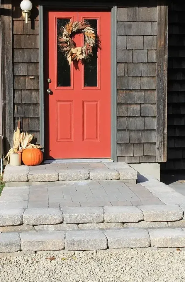 A vibrant red front door with a seasonal wreath, accented by a pumpkin and corn husks on stone steps of a wooden home.
