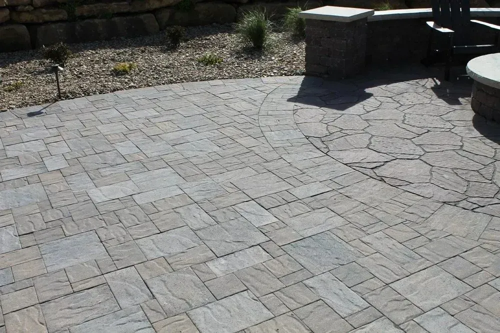 A gray, rectangular paver patio features a circular inlay design, bordered by a stone retaining wall and landscaped area.