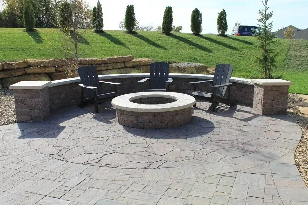 A stone fire pit with three dark chairs on a paved patio, set against a low retaining wall and a grassy hill.