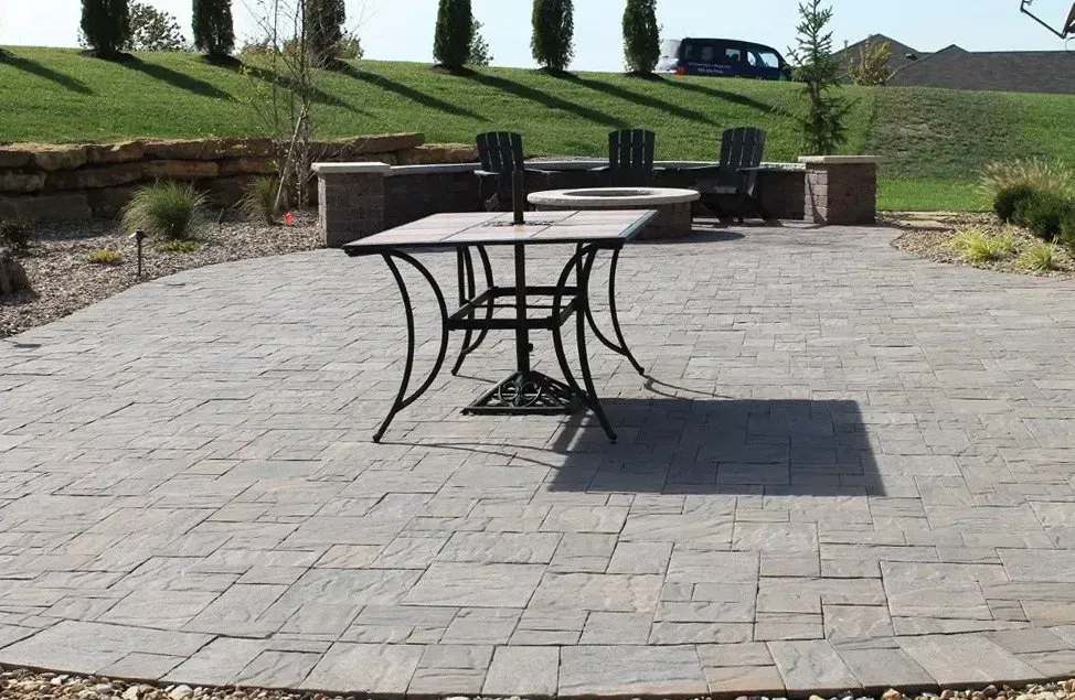 A stone patio features a square metal table and chairs, set against a stone retaining wall and a grassy, tiered landscape.