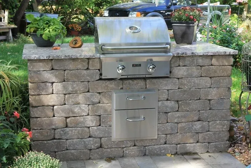 A stone-built outdoor kitchen island with a stainless steel built-in grill and storage drawers, set in a backyard.