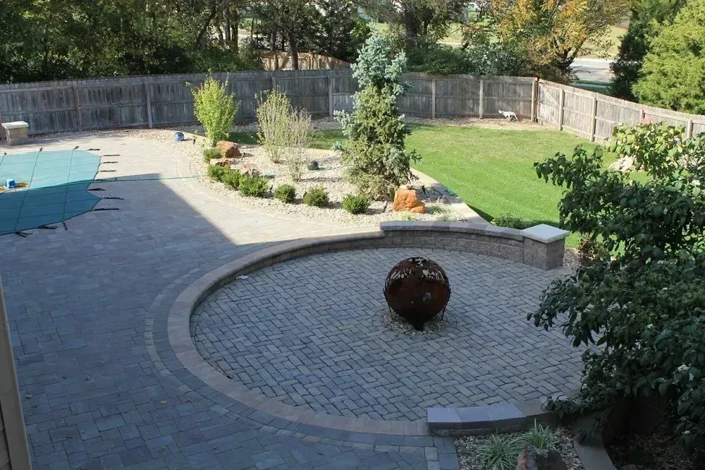 A backyard patio featuring a circular stone fire pit area, a swimming pool, landscaping, and a fenced-in grass lawn.