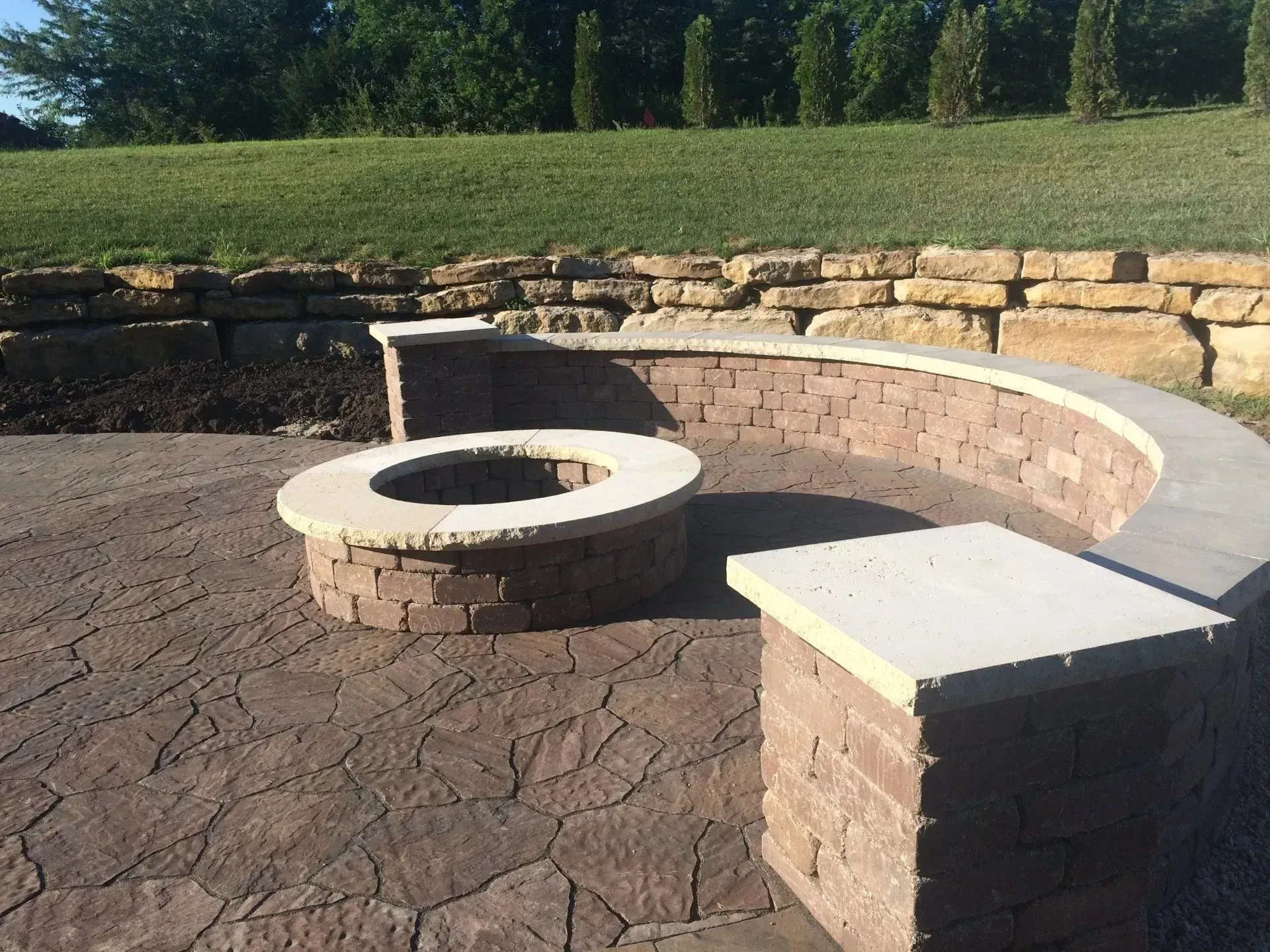 A circular stone fire pit sits on a paved patio with a curved stone seating wall in an outdoor landscape.