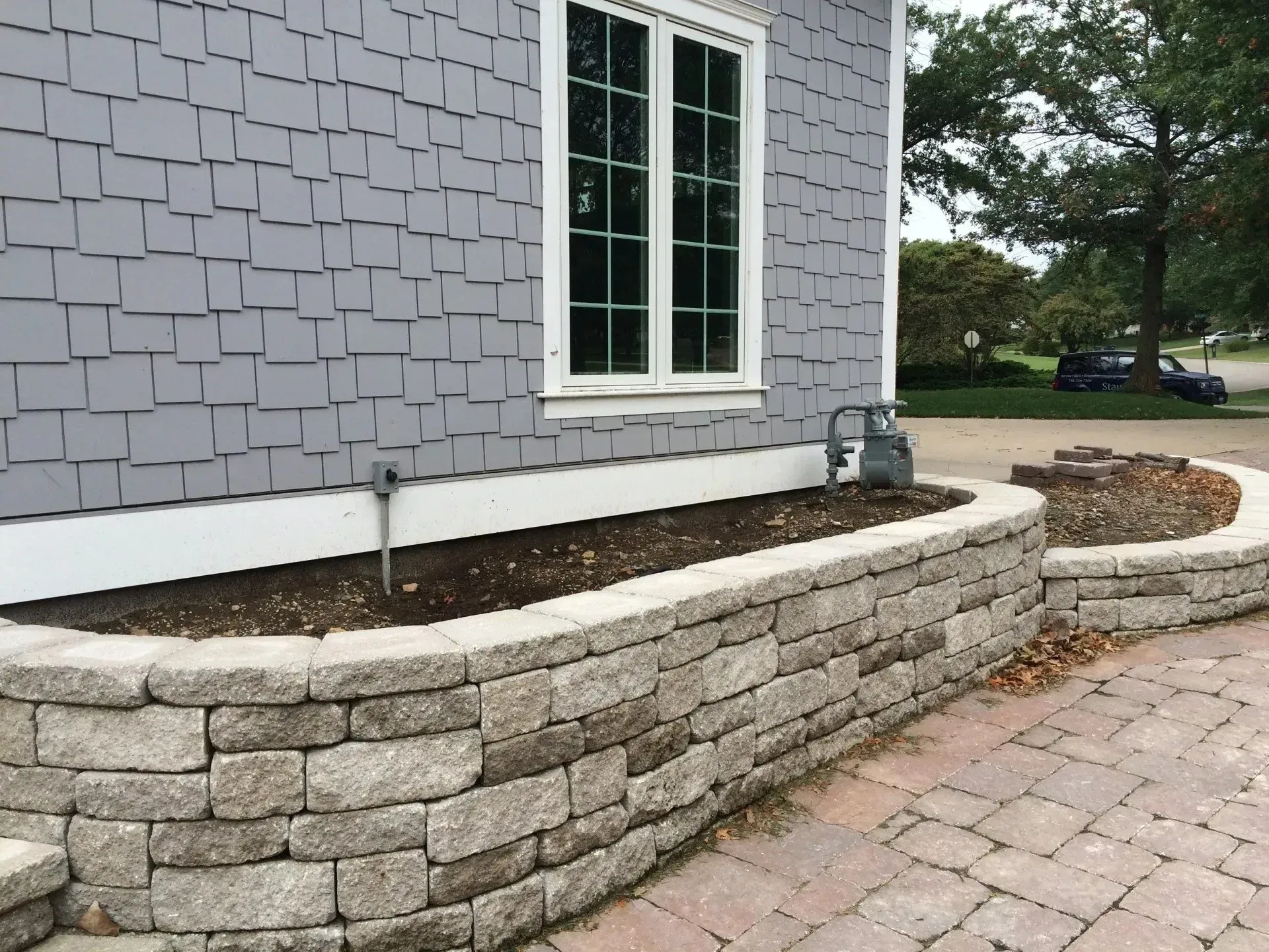 A curved stone retaining wall filled with dark soil sits in front of a gray shingled house wall with a white window.