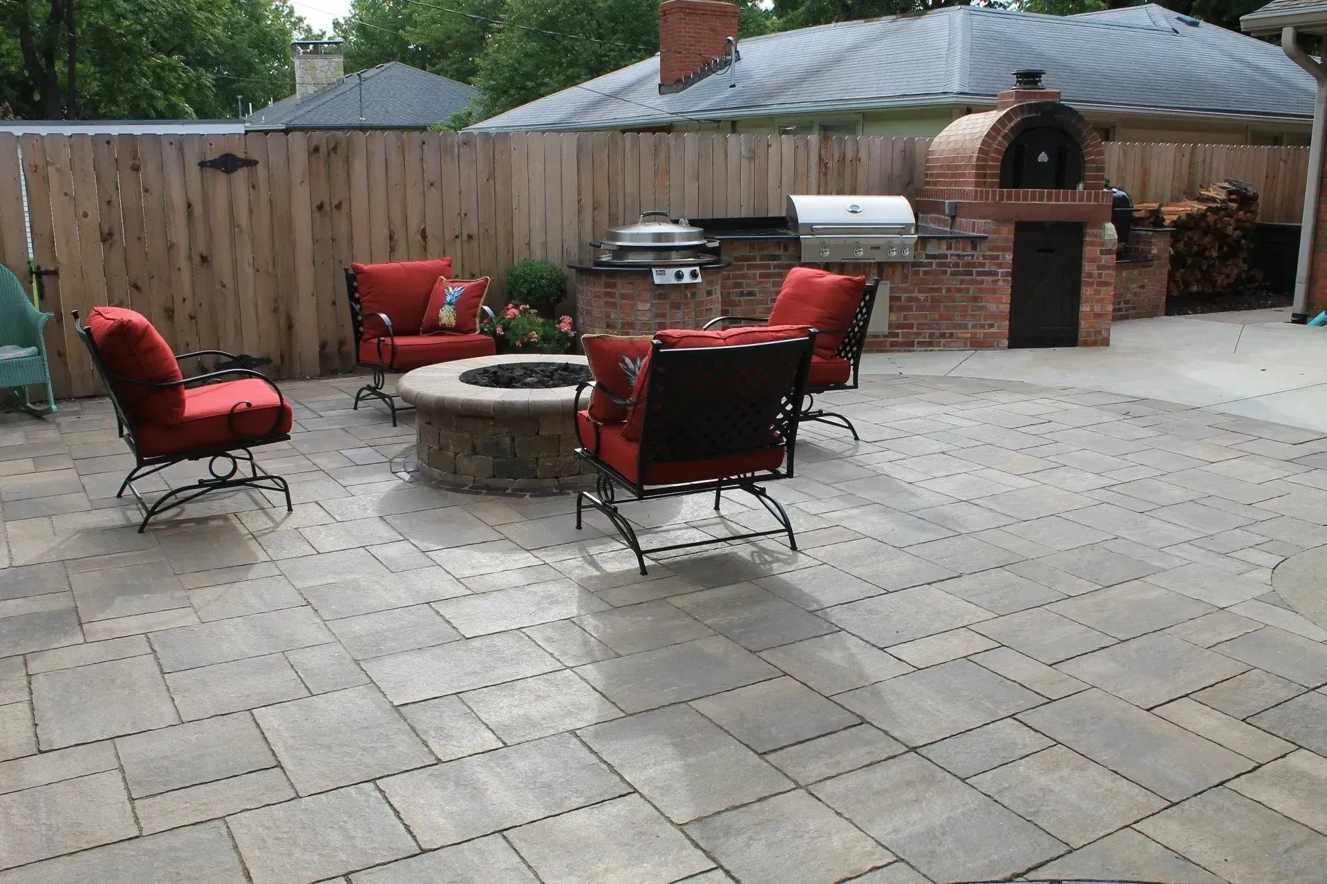 A backyard patio featuring a stone fire pit, four red cushioned chairs, a brick outdoor kitchen, and a pizza oven.