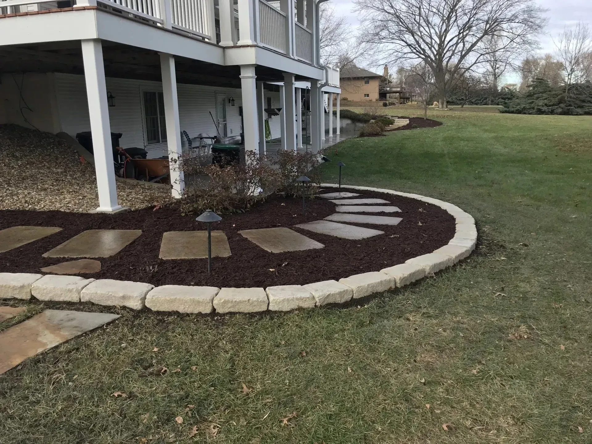 A curved landscape bed with stone pavers, dark mulch, and beige stone edging beneath a house deck and lawn.