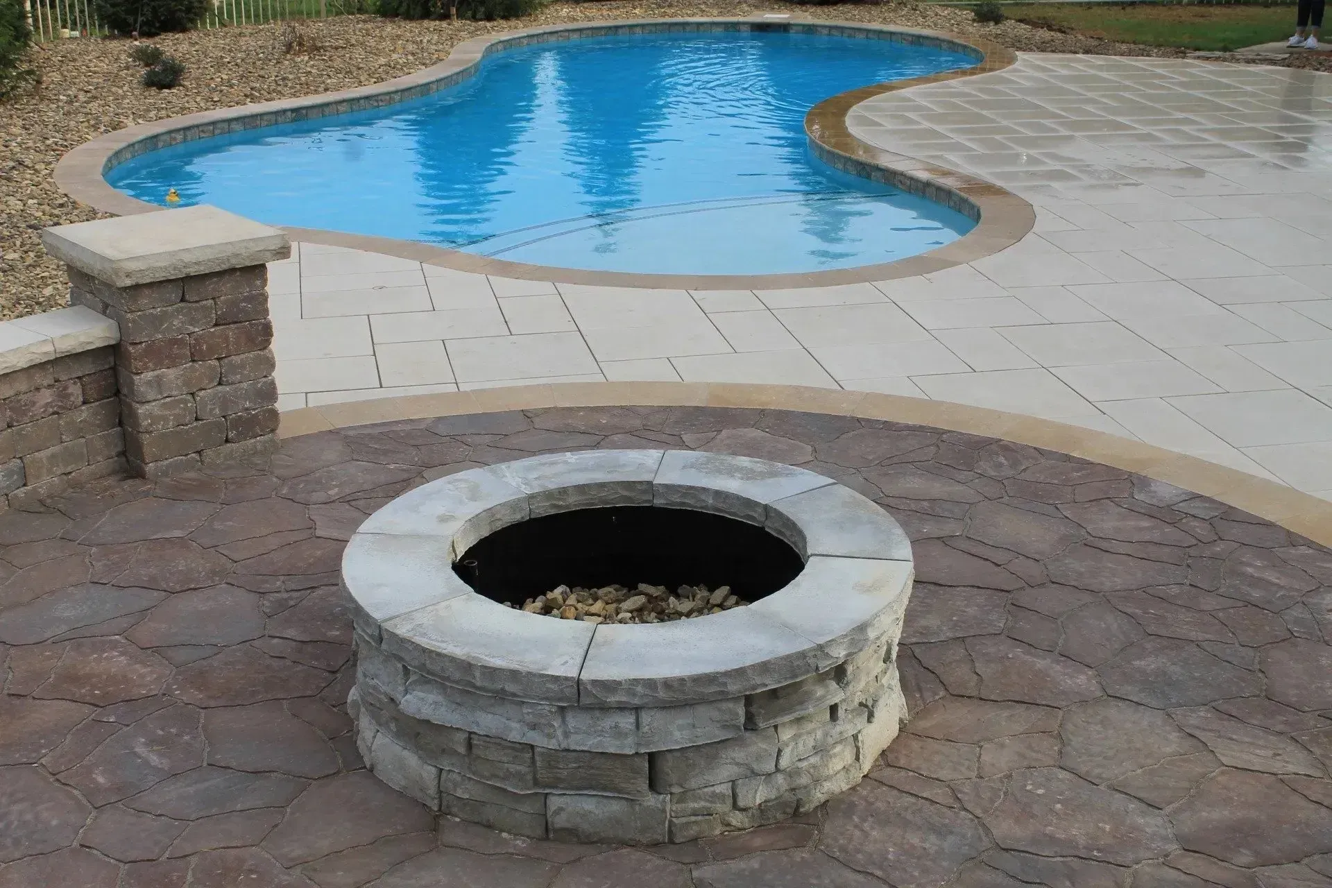 A stone fire pit sits on a brown stamped concrete patio next to a light-colored paved deck and a kidney-shaped pool.