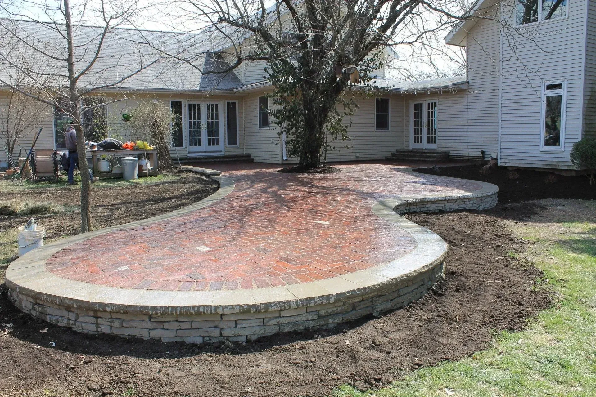 A curved brick patio with stone borders sits in a backyard next to a house with white siding and a large tree.