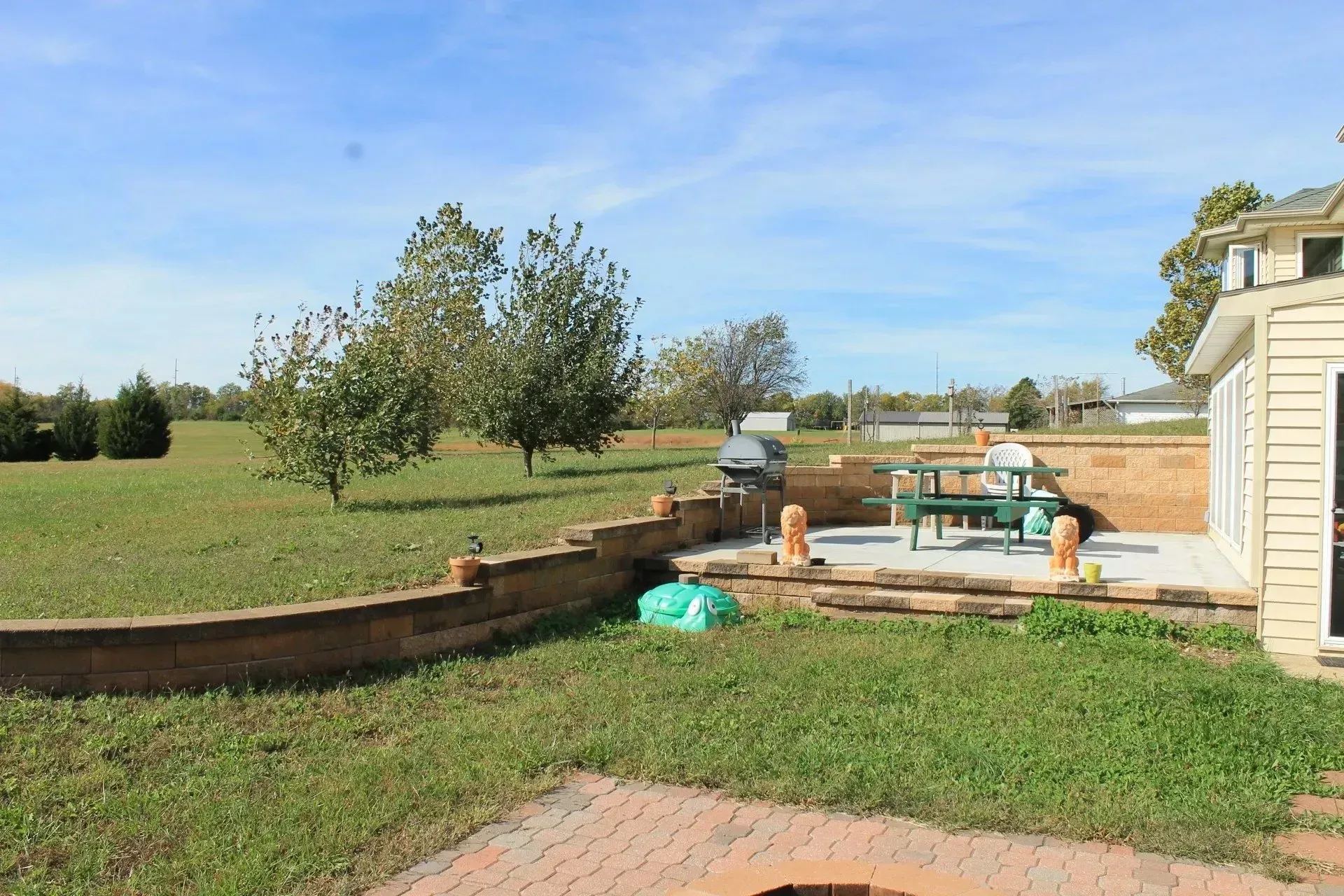 A backyard patio with a stone wall, grill, and picnic table overlooking a grassy lawn with trees under a blue sky.