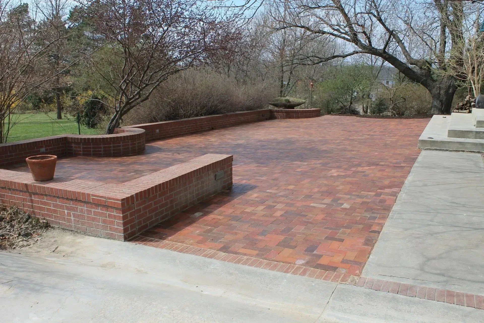 A red brick patio with low surrounding brick walls, set against a backdrop of trees and a concrete walkway.