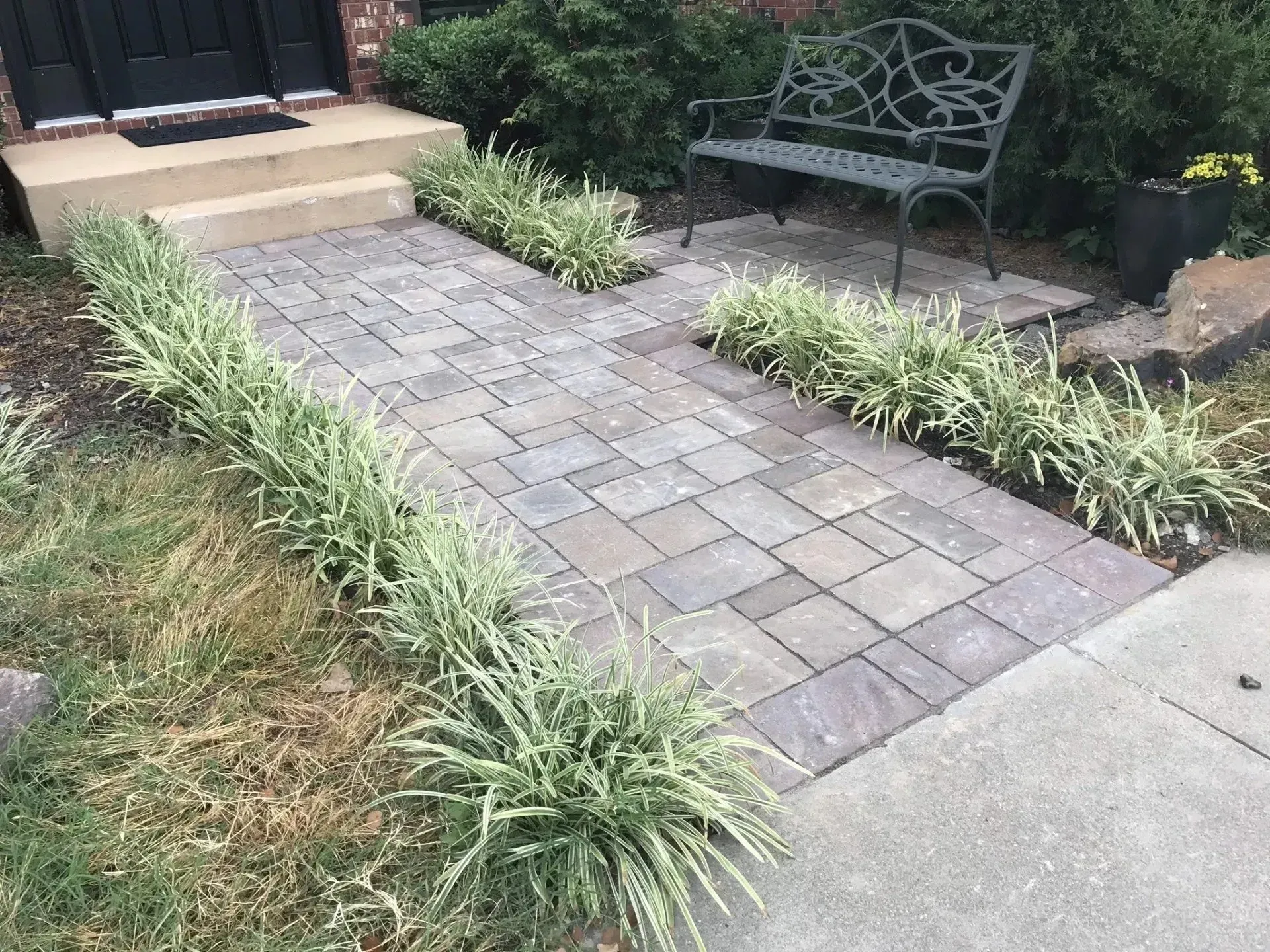 A gray paver walkway leads to a house entrance with an ornate metal bench tucked into a small patio area on the right.