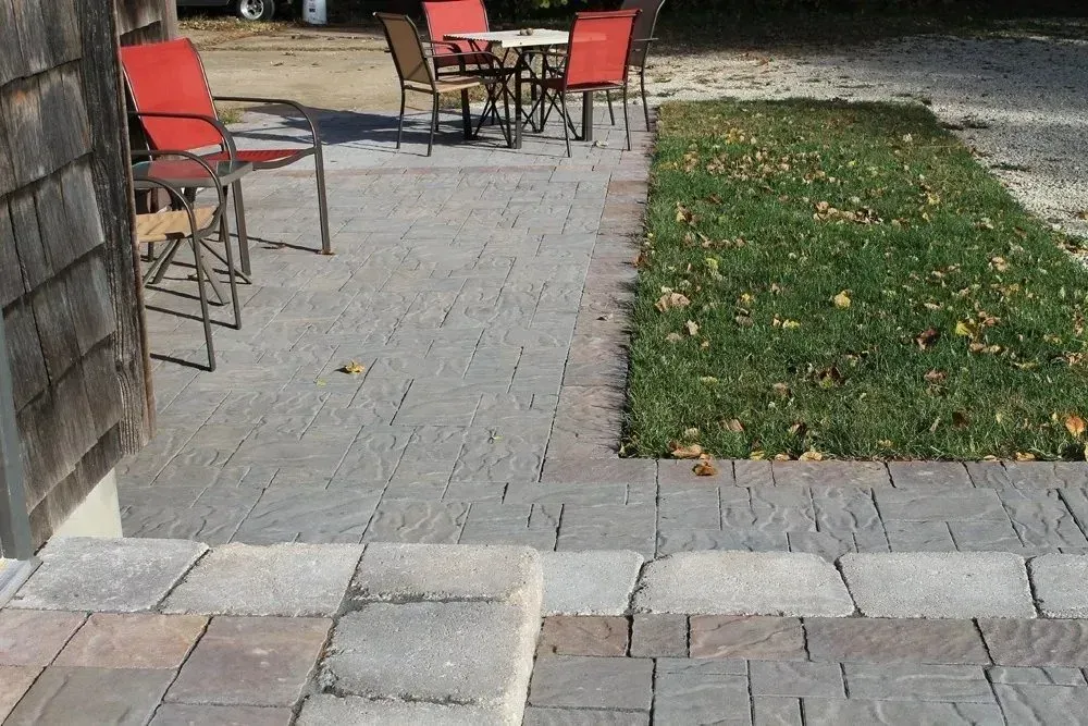 A stone-paved patio with several red chairs and a small table set outdoors next to a grassy lawn.