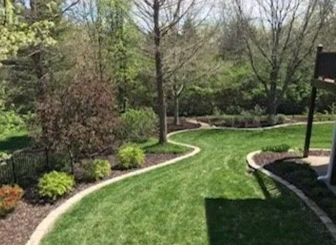 A curved, manicured grass lawn bordered by tan stone edging, surrounded by mulch beds with shrubs and trees.
