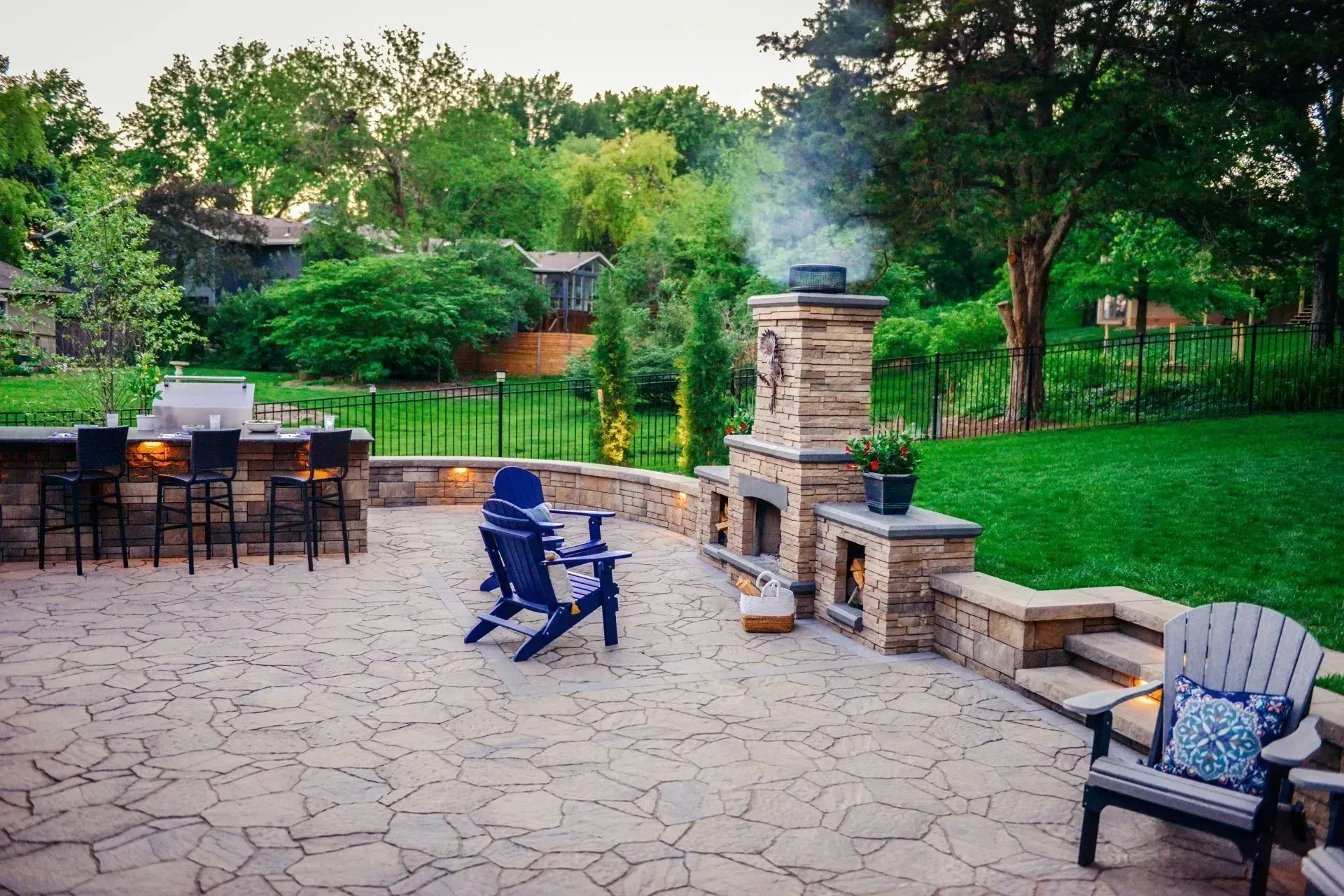 A stone patio at dusk features an outdoor fireplace, a kitchen counter with bar stools, and casual lawn chairs.