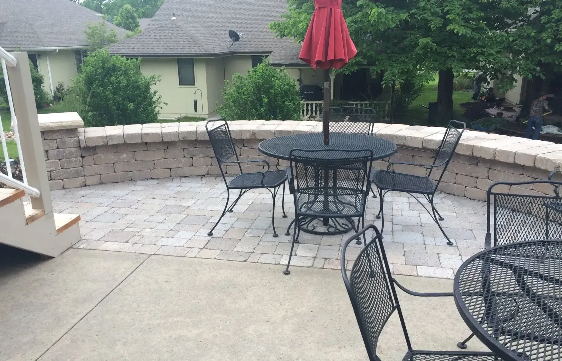 A patio with a stone wall, a table with a red umbrella, and several metal chairs on a sunny day.