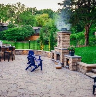 A stone patio with a fireplace, two blue chairs, a bar counter, and green backyard landscaping.