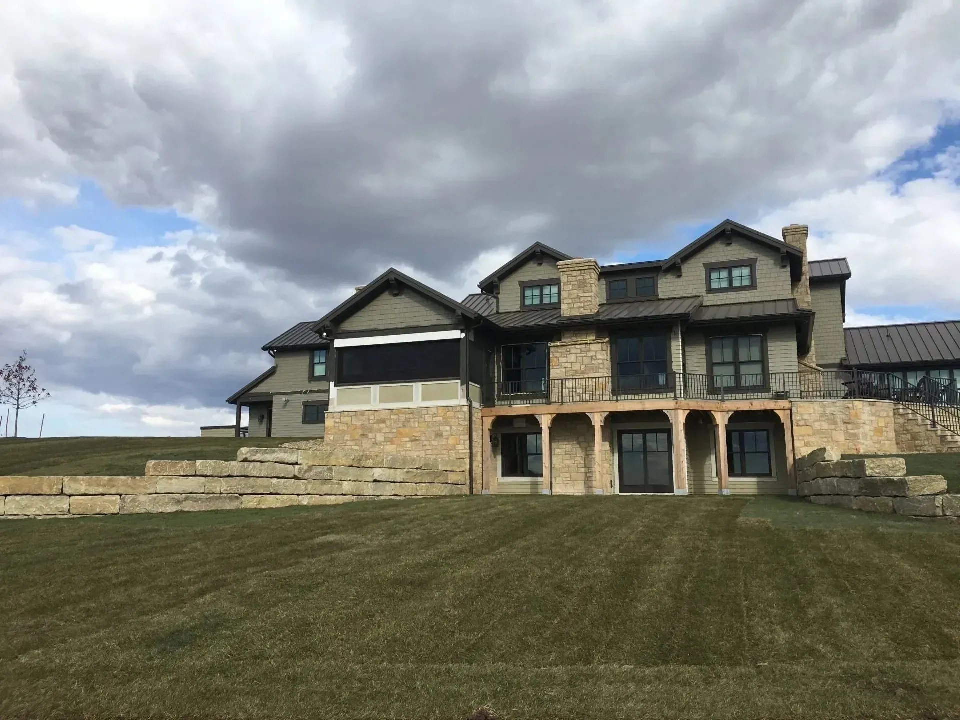 Large, multi-story stone and gray siding house with multiple gables and a terraced lawn under a cloudy sky.