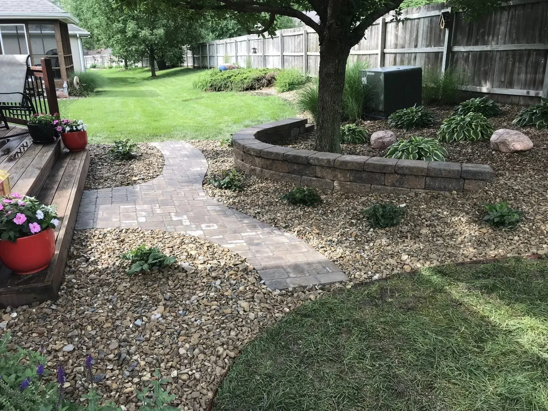 A paver path winds through a backyard garden with a stone retaining wall encircling a tree, next to a wooden deck.