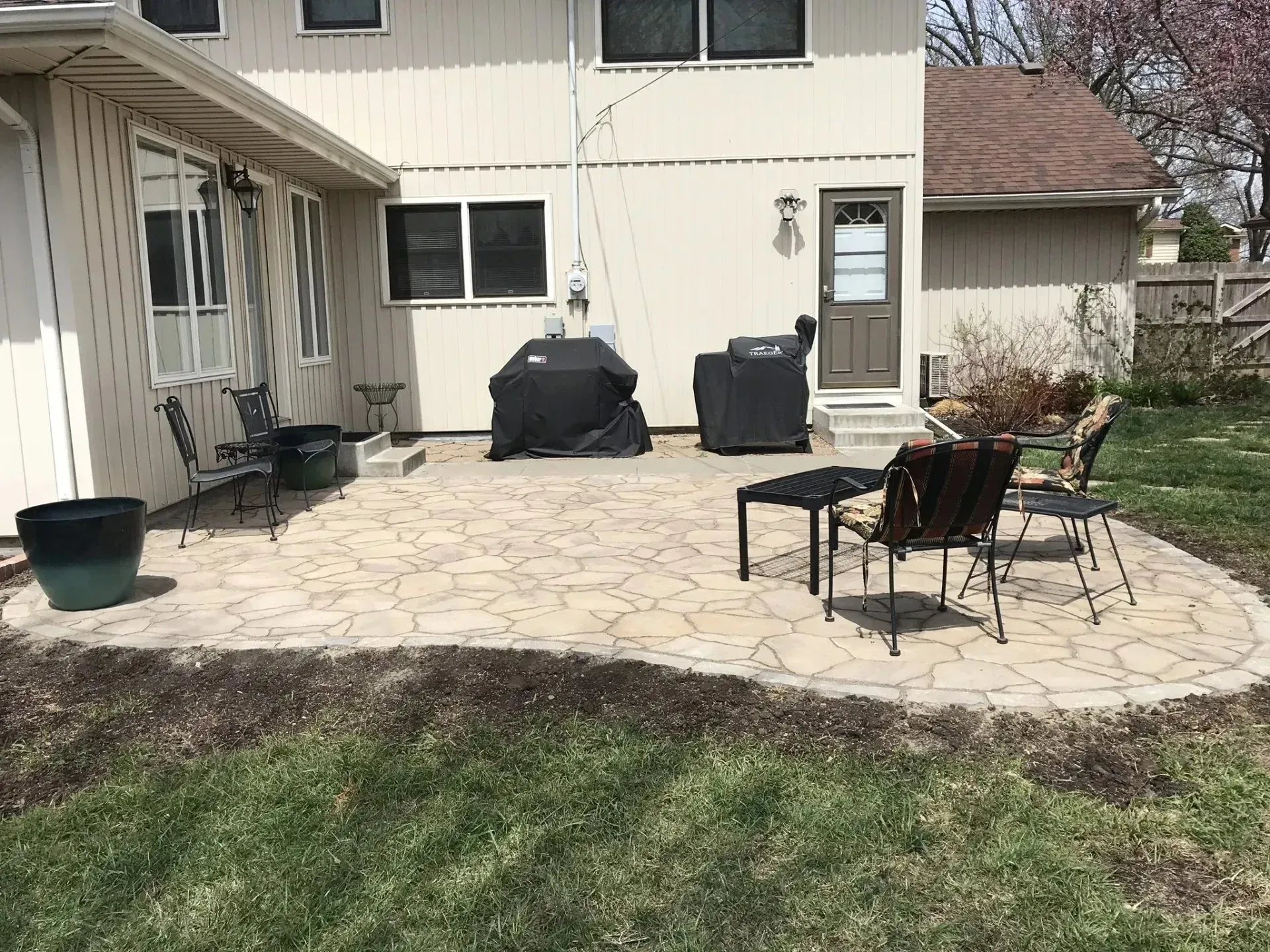 A light-colored house exterior with a patterned stone patio, outdoor furniture, and covered grills in a grassy backyard.