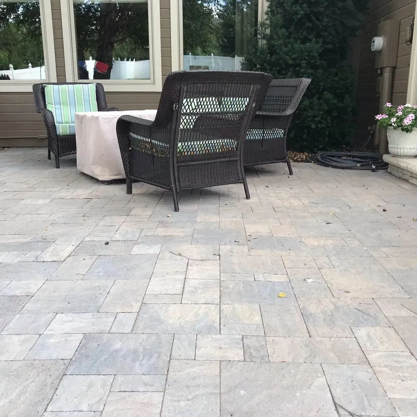 Wicker patio furniture with patterned cushions arranged on a stone paver patio next to a house with large windows.