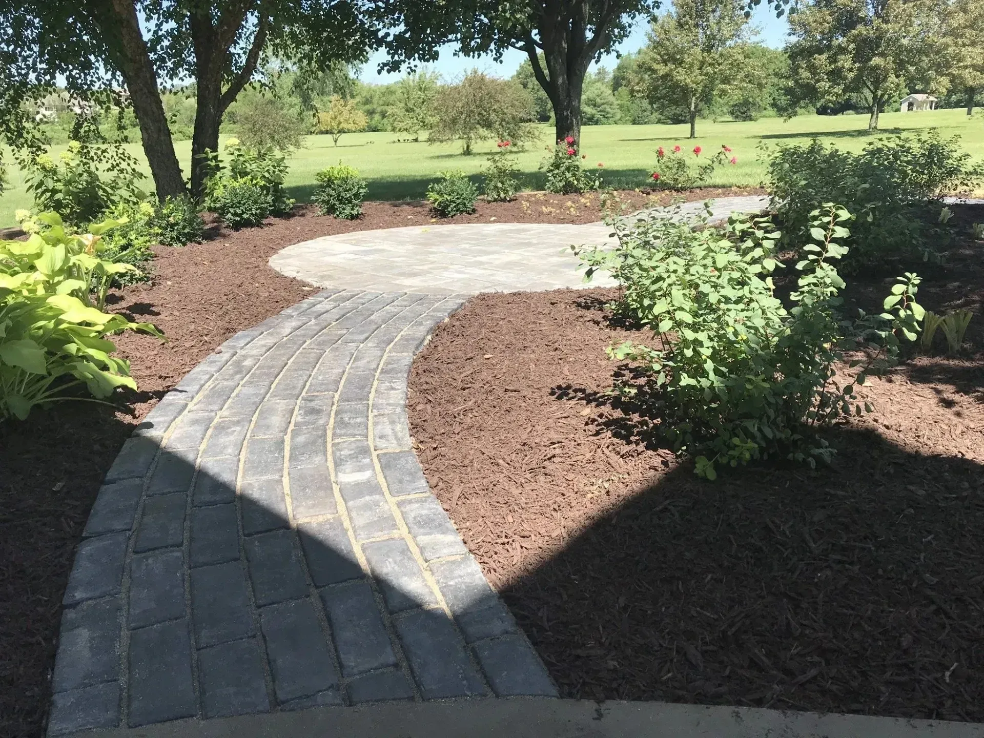 A gray paver path curves through a mulched garden bed leading to a circular stone patio under trees on a sunny day.