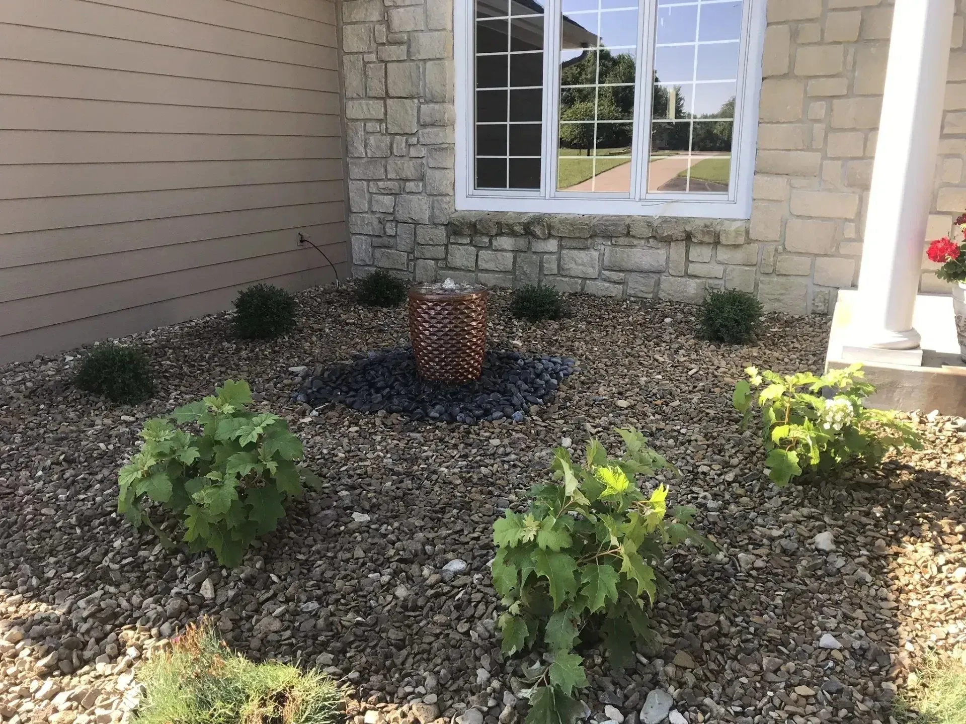 A decorative fountain centered in a landscaped yard with stone mulch and green bushes in front of a stone house wall.