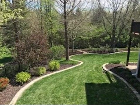 A grassy backyard featuring curved stone-edged garden beds, mulch, small shrubs, and trees under a clear sky.