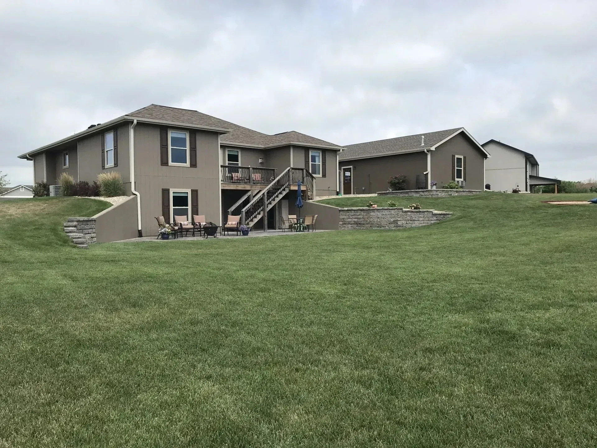A tan, two-story house with a wooden deck and basement patio sits on a large, grassy lawn under a cloudy sky.