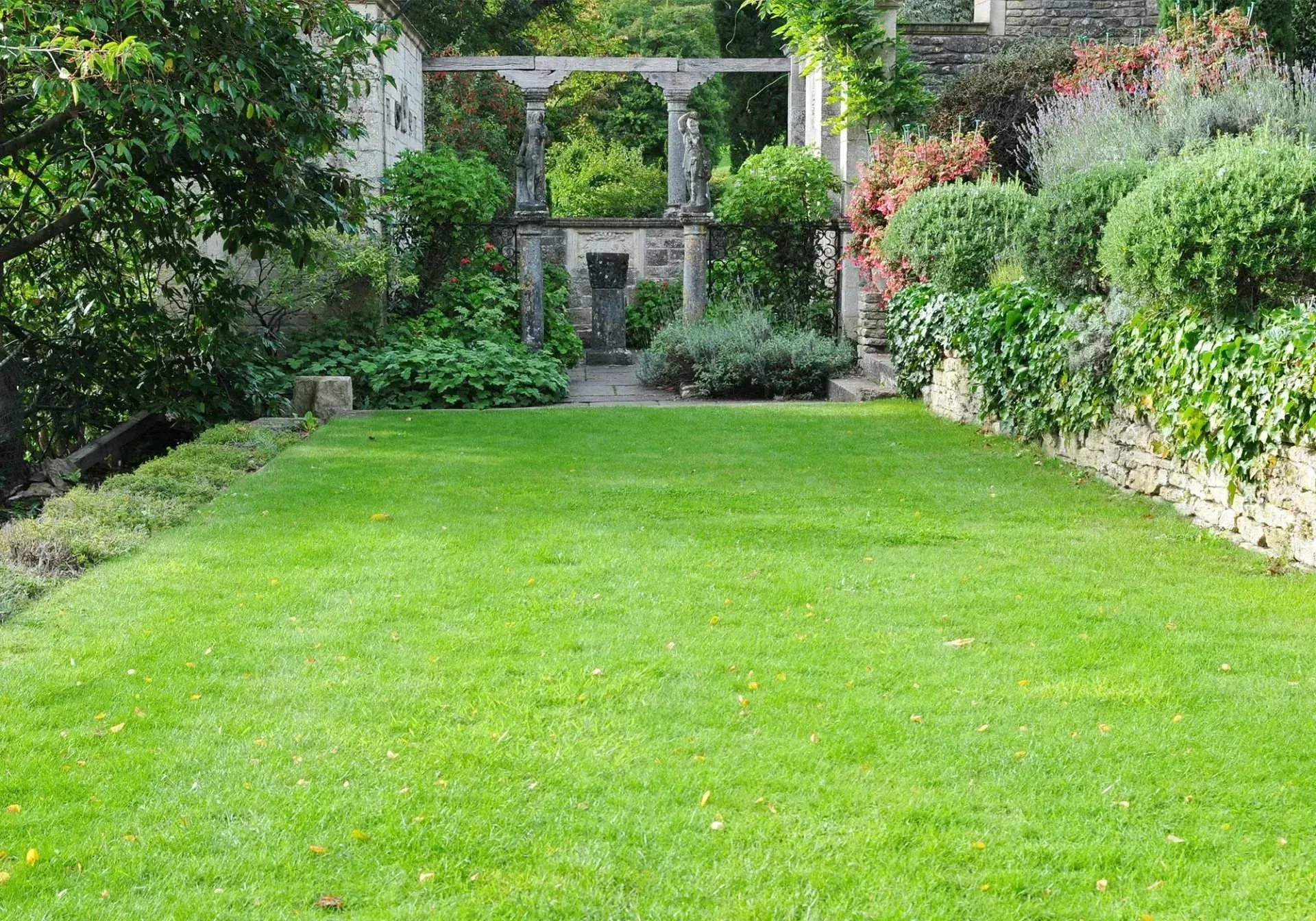 A bright green lawn leads toward a stone pergola in a lush, tiered garden with shrubs and trees.