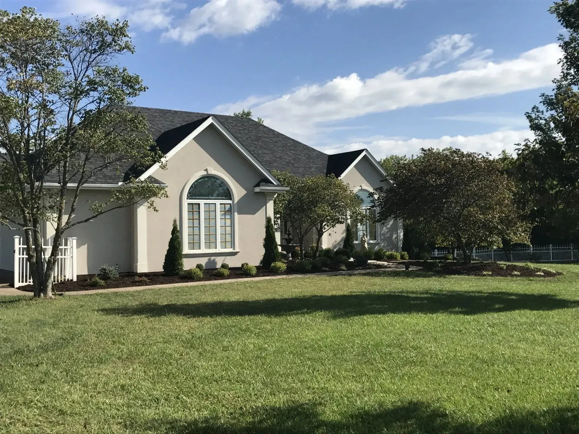 A light-colored, single-story suburban home with a dark shingled roof, arched front window, and a large front lawn.