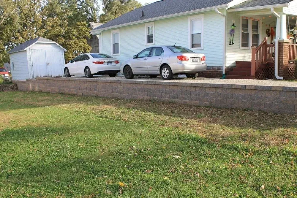 A light blue house with two parked sedans sits behind a stone retaining wall next to a small white shed.