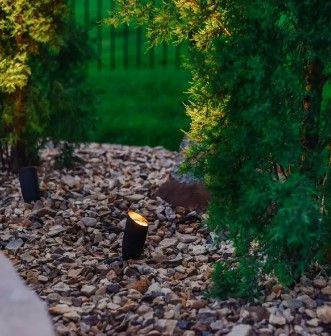 Small landscape lights placed in a bed of gravel between evergreen bushes at dusk.