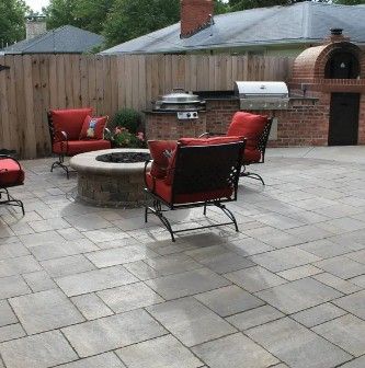 A backyard patio with light-colored pavers, red cushioned chairs around a circular fire pit, a grill, and brick oven.