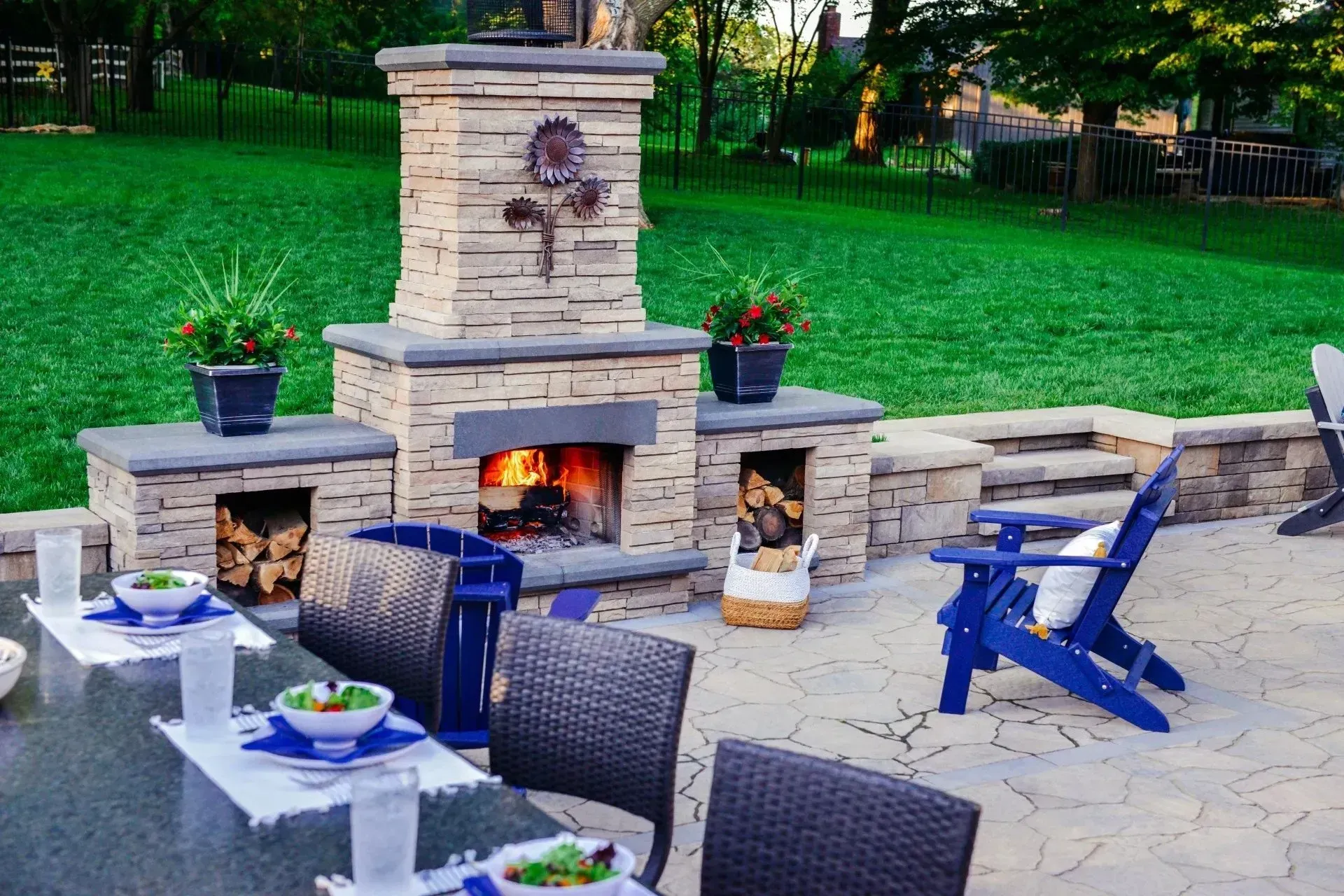 A stone outdoor fireplace with an active fire and side log storage on a backyard patio with a dining table and chairs.