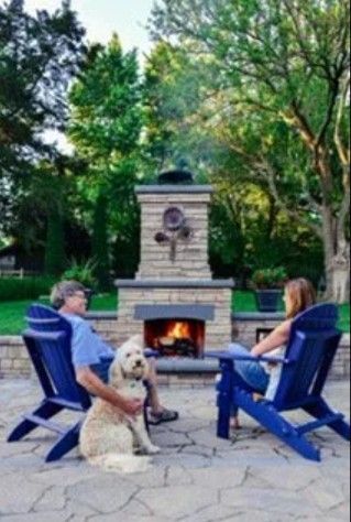 A person and their dog sitting on blue chairs in front of an outdoor stone fireplace in a wooded backyard.