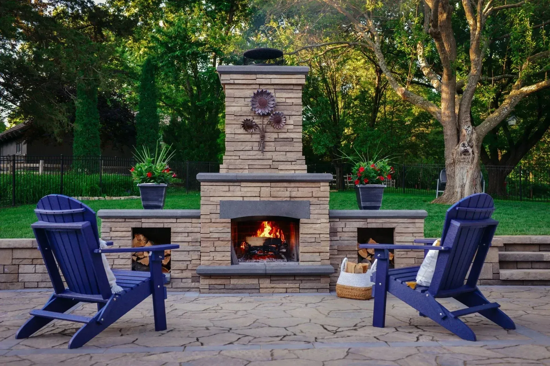 Two blue Adirondack chairs face a stone outdoor fireplace with a burning fire, set on a patio in a lush backyard.