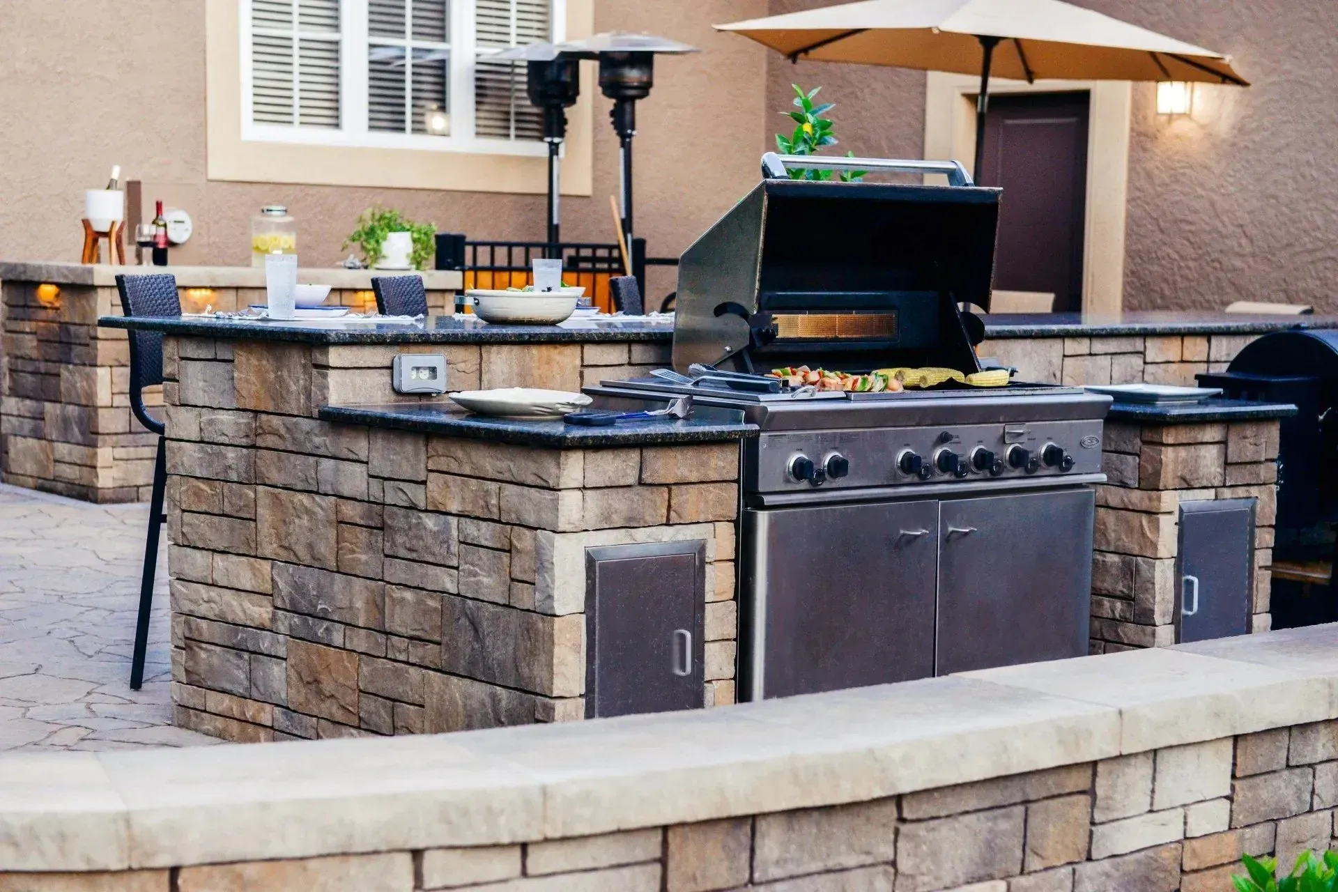 Outdoor stone kitchen island with a built-in stainless steel grill and a large umbrella on a patio.