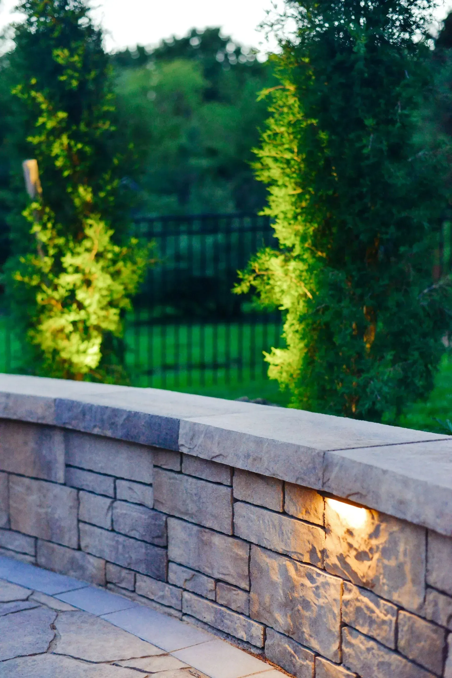 A stone retaining wall with an integrated light fixture, set against tall green trees and a dark metal fence at dusk.