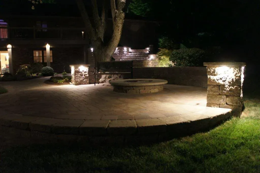 A stone patio at night features a fire pit, bench, and glowing stone pillars, with a house visible in the background.