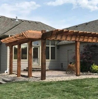 A wooden pergola attached to the back of a light-gray house, overlooking a stone patio and grass lawn.