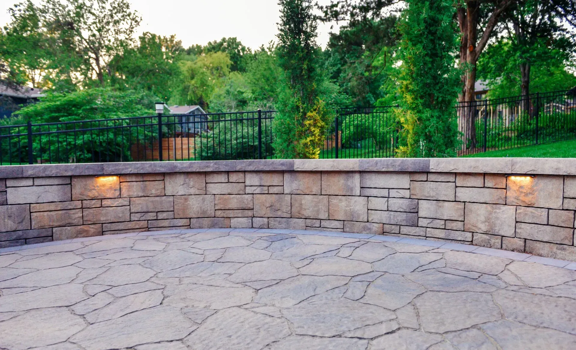 A curved stone retaining wall with built-in lights, surrounding a flagstone patio against a backdrop of trees and a fence.