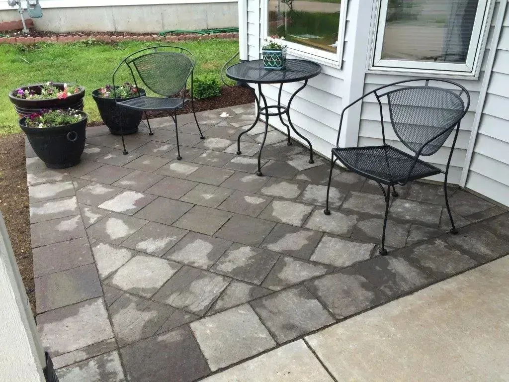 A small patio with two metal chairs, a matching bistro table, and two dark planters with flowers on a stone paver floor.