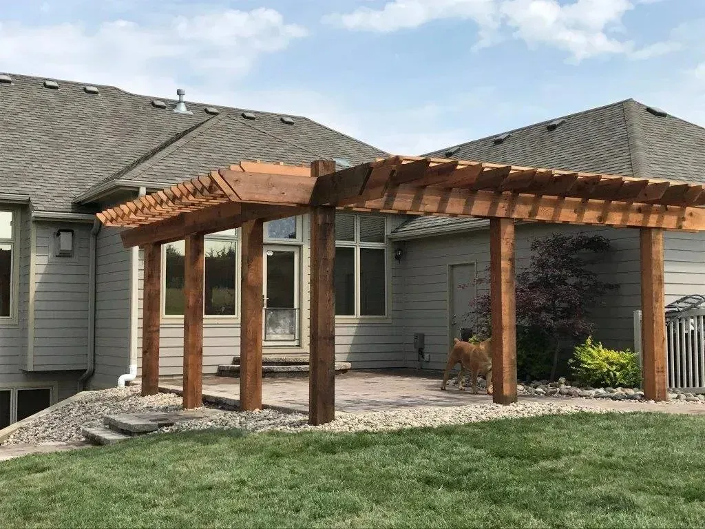 A wood pergola stands over a stone patio against the rear wall of a beige house with a grey shingled roof.
