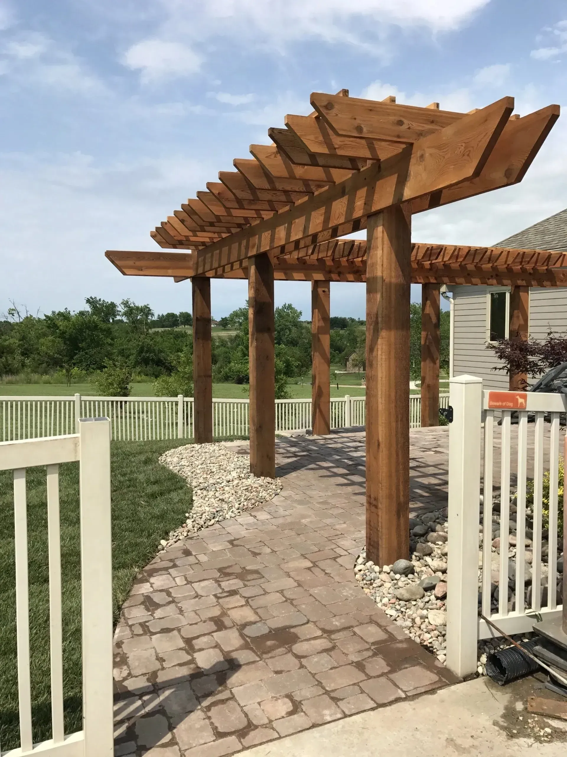 A wooden pergola stands over a stone paver path next to a white fence, with green trees and a field in the background.