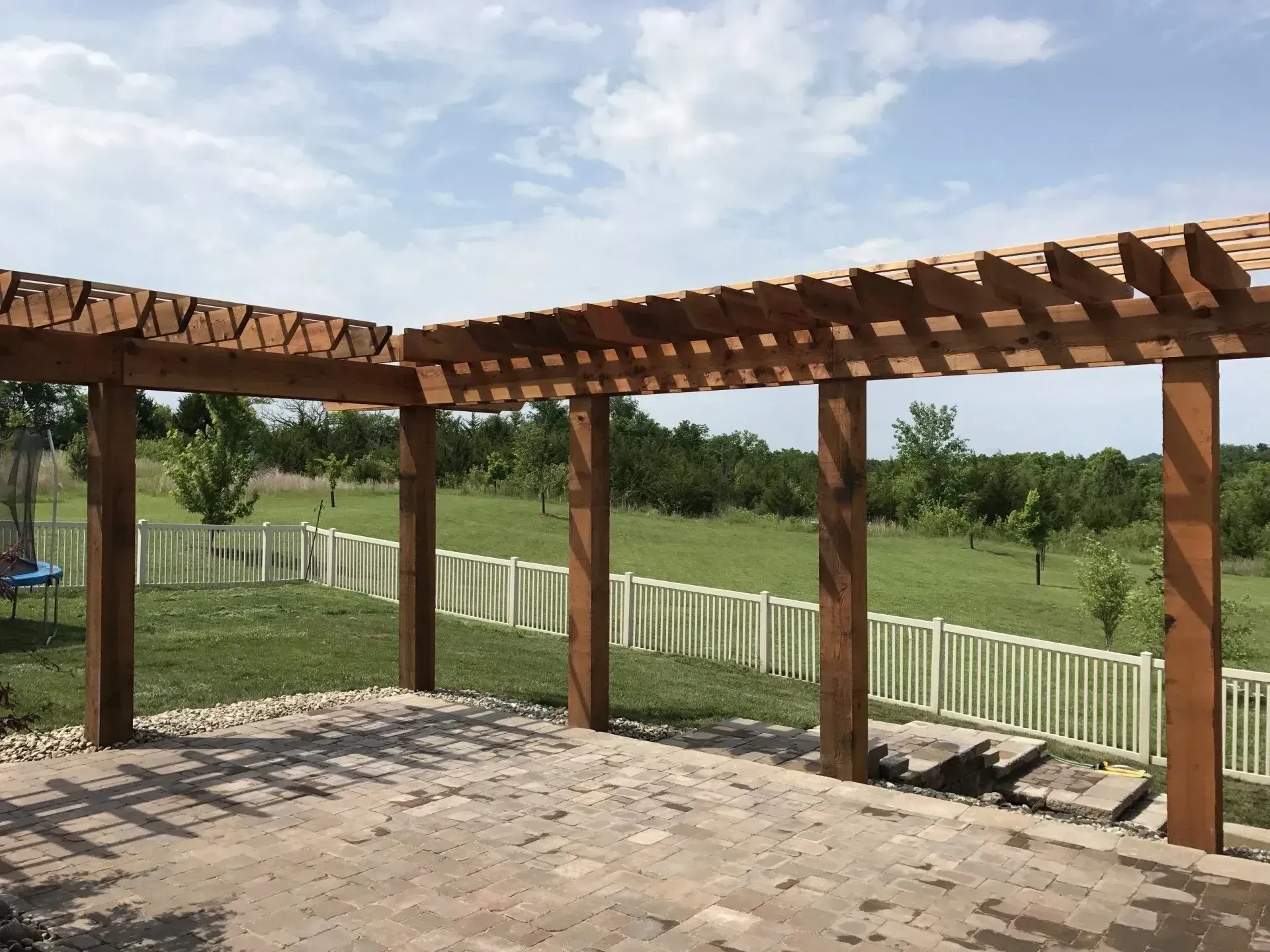 Wooden pergola over a stone patio in a backyard with a green lawn and a white fence in the background.