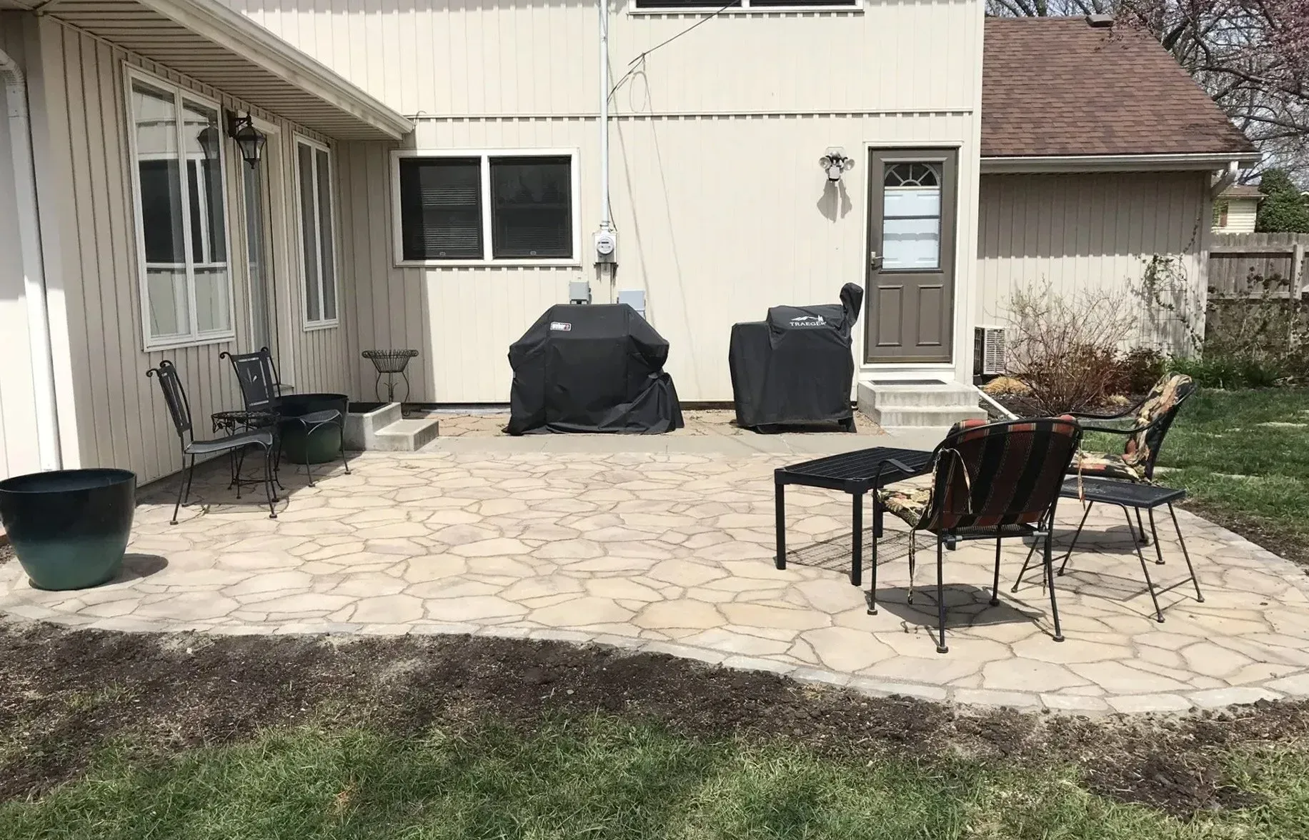 A tan-colored stone patio with outdoor chairs, a table, and two black-covered grills by the side of a house.