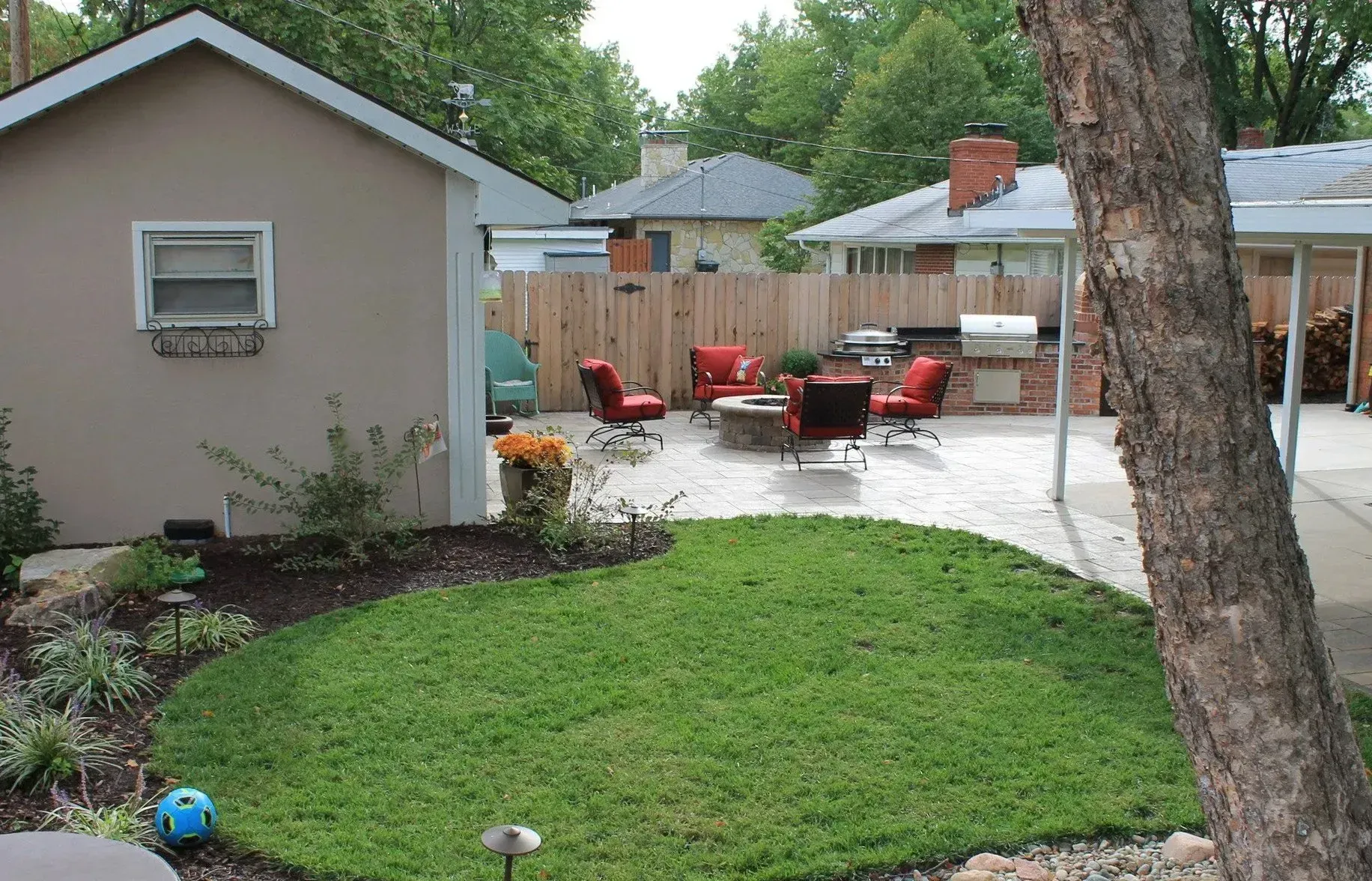 A backyard featuring a circular lawn, paved patio with outdoor furniture, a brick fire pit, and a small tan shed.
