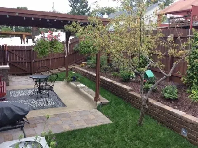 A backyard patio with a pergola, stone pavers, a small table and chairs, a retaining wall, and a tree in a garden bed.