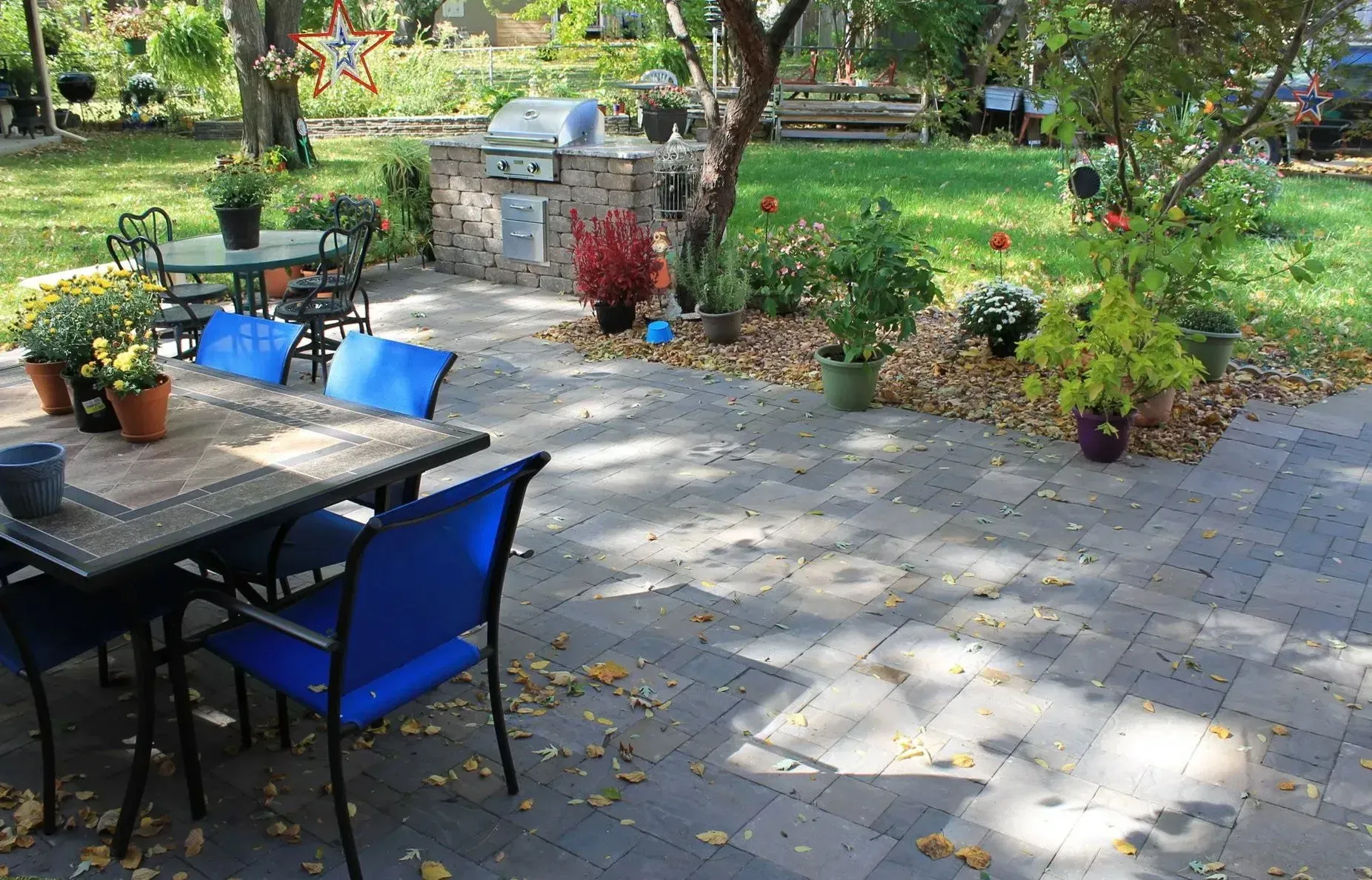 A backyard patio with a stone grill, dining sets with blue chairs, and potted plants on a paved stone surface.