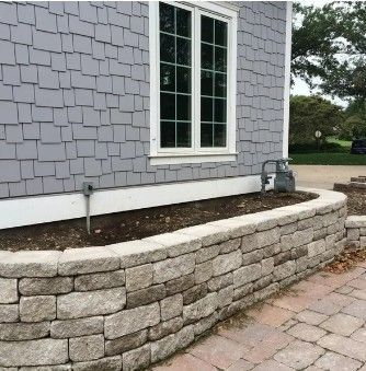 A low, curved stone retaining wall sits in front of a grey-shingled house with a white-framed window and a gas meter.
